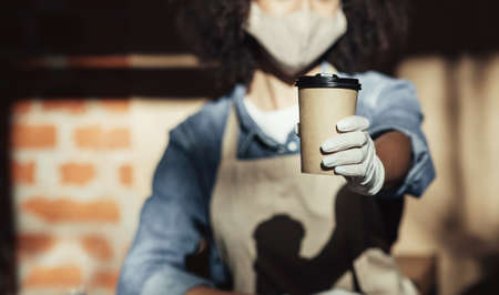 Barista gives online order of coffee to go during covid-19 quarantine and social distancing. Young african american female waiter in apron, face mask and gloves holds drink in modern cafe, croppedの写真素材