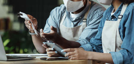 Finance, small business, COVID-19 pandemic. Sad millennial african american couple owners of small business in protective masks work with laptop and smartphone and accounts in cafe, cropped, panoramaの写真素材