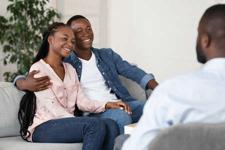 Marriage Reconciling. Happy African American Spouses Sitting At Marital Counselors Office After Successful Therapy, Young Black Lovers Smiling And Embracing On Couch At Psychotherapist, Free Spaceの写真素材