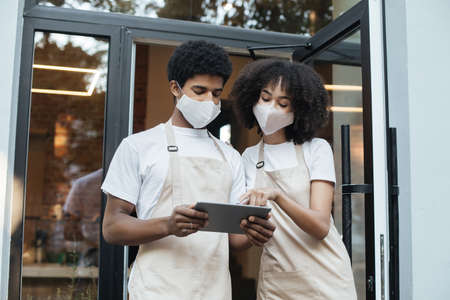 Outdoor service, acceptance online orders and new normal during covid-19 quarantine. Millennial african american guy and woman in aprons and protective masks use tablet near front door, free spaceの写真素材