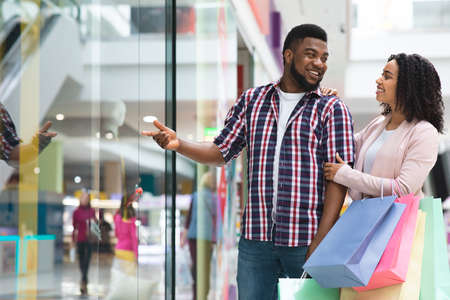 Happy Black Man Shopping In Mall With Girlfriend, Pointing At New Clothing In Showcase, Showing Good Price Offer Discount, Young African American Couple Enjoying Seasonal Sales, Copy Spaceの写真素材