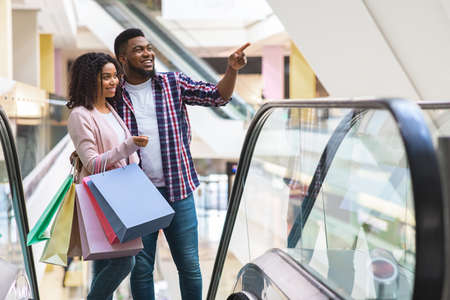 Portrait Of Happy African American Couple At Escalator In Department Store, Young Black Spouses Shopping Together In Mall, Enjoying Making Purchases, Carrying Lots Of Bright Shopper Bags, Free Spaceの写真素材