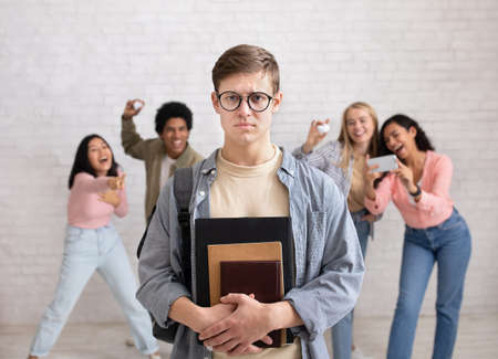 Humiliation and bullying at school. Focus on sad young guy in glasses and notebooks, and multiracial students filming video and throwing papers at guy and laugh at pupil, free space, studio shotの写真素材