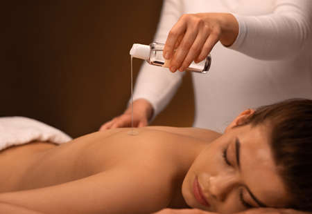 Closeup of masseuse pouring oil on sleeping woman back at luxury spa, body care, massage, spa concept. Young attractive lady enjoying relaxing massage with aromatherapy, copy spaceの写真素材