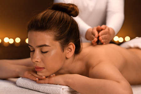 Closeup of beautiful woman enjoying back massage at luxury spa decorated with candles. Peaceful young lady laying on massage table, having healing session at romantic atmosphereの写真素材