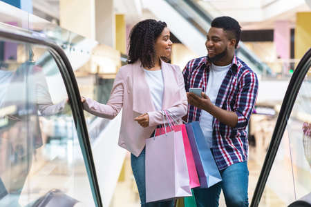 First Meet. Handsome Black Man Introducing With Beautiful Woman In Shopping Center, Asking Her Phone Number While Riding Escalator Together In Department Store, Woman Smiling In Return, Free Spaceの写真素材