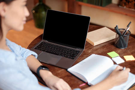 Online Course Concept. Over the shoulder view of female student sitting at table and writing in notebook, using laptop with black empty screen for mock up template, watching lecture or webinarの写真素材
