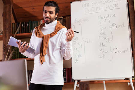 Distance Education And E-Teaching. Young Indian Teacher Teaching Online Standing Near Blackboard With English Language Grammar Rules Having Webinar Via Video Call And Laptop At Home. Remote Studyの写真素材