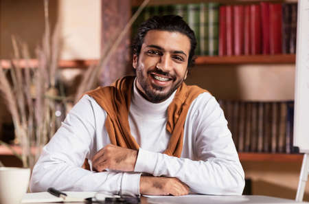 Teaching. Portrait Of Young Indian Teacher Man Smiling To Camera Sitting At Workplace Over Books Shelf Background Indoors. Modern Education, Tutoring Conceptの写真素材