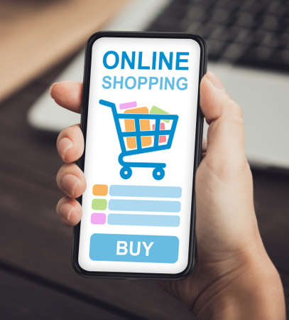 Unrecognizable Lady Doing Online Shopping On Smartphone Sitting At Desk Indoors. Female Hand With Cellphone And Opened Shop Application. Cropped, Vertical, Closeupの写真素材
