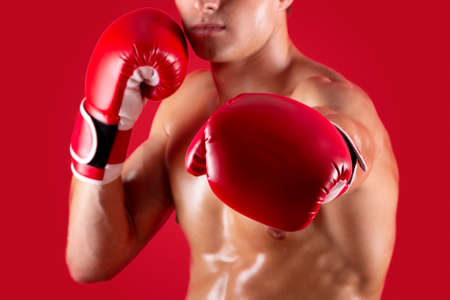 Determined young boxer in fighting gloves making punch at camera on red studio background, selective...