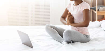 Unrecognizable black woman making morning exercise in front of laptop at home, sitting on bed and...