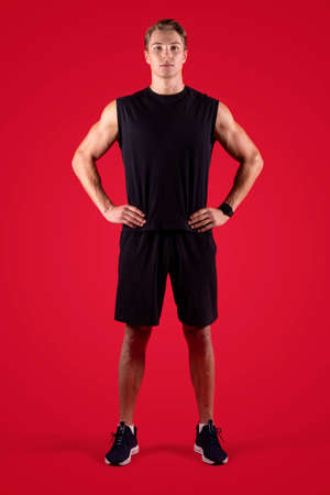 Full length portrait of determined male fitness model looking at camera on red studio background. Serious millennial athlete in black sportswear posing with hands on hips. Professional sports conceptの写真素材