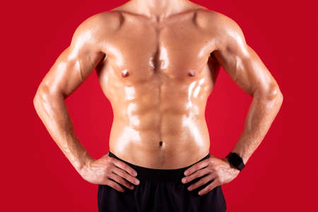 Cropped view of handsome young sportsman with bare torso posing with hands on hips over red studio...