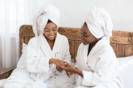 Young black woman happy to see her girlfriends engagement ring, drinking champagne together, bedroom interior. Two joyful african ladies in bathrobes having bridal party at home, celebrating affianceの写真素材