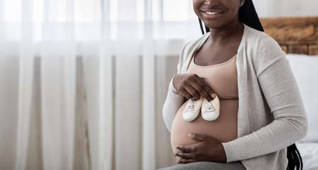 Happy Expectation. Black Expectant Woman Holding Small Baby Shoes Near Her Pregnant Belly, Happy African Future Mother Enjoying Maternity Time At Home, Cropped Image With Copy Space, Panoramaの写真素材