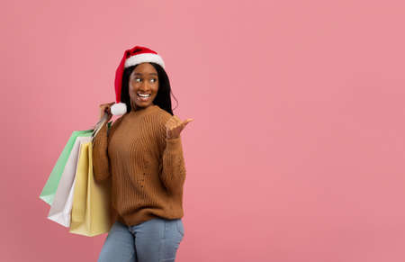 Huge Christmas sale. Attractive young black woman in Santa hat holding gift bags and pointing aside at blank space, pink studio background. African American lady shopping for Xmasの写真素材