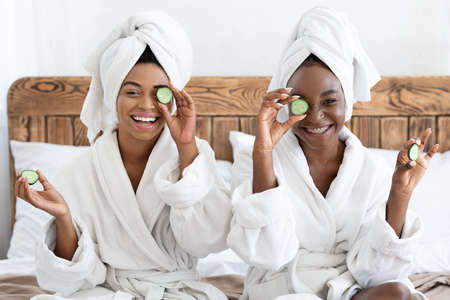 Two cheerful black ladies in bathrobes holding cucumber slices, using fresh vegetables in face care...