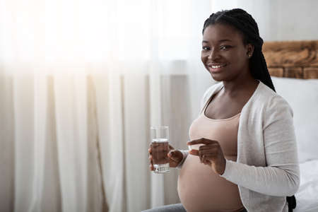 Prenatal Vitamins. Pregnant Black Woman Holding Supplement Pill And Glass Of Water, Taking Healthy Nutrients, Enjoying Maternity, Sitting On Bed At Home, Looking At Camera, Copy Spaceの写真素材