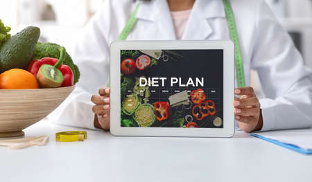 Black dietologist doctor lady demonstrating digital tablet with opened diet nutrition plan app with...