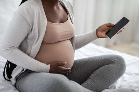 Unrecognizable African American Expectant Mother Using Smartphone With Black Screen At Home, Browsing Pregnancy App Or Shopping Online While Relaxing On Bed, Touching Her Pregnant Belly, Cropped Imageの写真素材