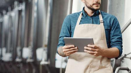 Modern plant management, mobile app and small business. Busy attractive man worker or owner in apron with digital tablet controls equipment in factory with large boilers for brewing beer, croppedの写真素材