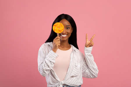 Young black woman covering her eye with bright lollipop and showing PEACE gesture on pink studio...