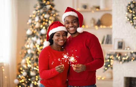 Happy New Year and Merry Xmas at home together. Smiling young african american guy and lady in Santa Claus hats hold sparklers, hug and look at camera in living room interior with tree and garlandsの写真素材