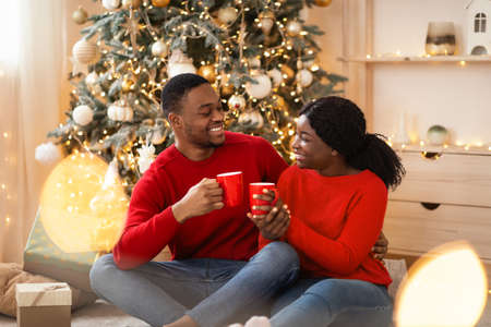 Romantic new year and christmas morning at home. Happy young african american guy and lady sit on floor and hold cups with hot drink in interior of living room with tree and garlands, free spaceの写真素材