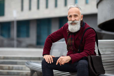 Handsome aged bearded man in stylish outfit with bag sitting on bench, smiling at camera, copy...