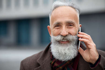 Closeup portrait of cheerful aged man in warm outfit talking on mobile phone while walking by...