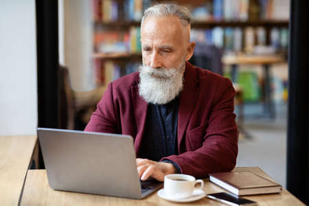 Aged businessman using laptop while having lunch break at cafe, drinking coffee, copy space. Elderly...