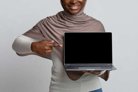 Mockup Image Of African Woman In Hijab Pointing At Laptop With Black Screen, Islamic Lady In Headscarf Demonstrating Online Offer Or New Website, Showing Copy Space For Your Design, Cropped Imageの写真素材