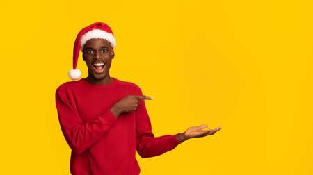 Excited Black Man In Santa Hat Pointing At Invisible Object On His Palm, Showing Christmas Gift Ideas, Standing On Yellow Background, Demonstrating And Advertising Something, Panorama With Copy Spaceの写真素材