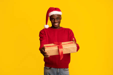 Cheerful black guy in santa hat and red sweater giving gift box at camera, presenting christmas present to somebody, positive african man enjoying winter holidays, isolated on yellow backgroundの写真素材
