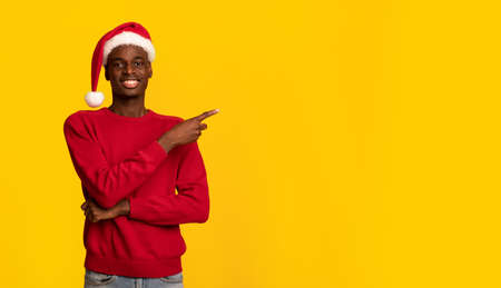 Check This. Cheerful Black Guy In Santa Hat Pointing At Copy Space Over Yellow Background With Index Finger, Demonstrating Empty Place For Your Text Or Design, Enjoying Christmas Sales And Offersの写真素材
