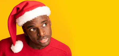 Interesting Offer. Curious African Guy In Santa Claus Hat Overhear And Looking Away At Copy Space On Yellow Background, Holiday Website Banner, Closeup Shot Of Interested Black Xmas Man, Panoramaの写真素材