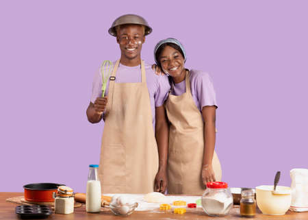 Portrait of happy black couple with dirty faces cooking together on violet studio background. Young...