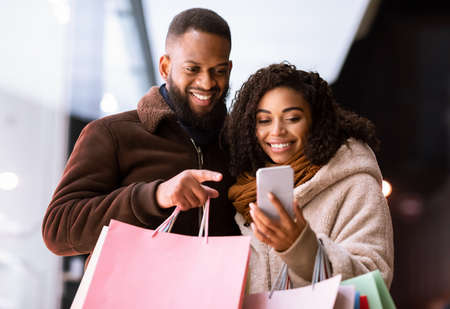 Shopping Together Concept. Portrait of happy african american couple using smartphone, pointing at gadget, holding shopping paper bags, standing outdoors in the evening. Retail, Sale, Technologyの写真素材