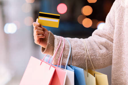 Easy Payment Concept. Closeup Of Unrecognizable Black Woman In Winter Coat Holding Credit Card And Bright Colorful Shopping Bags From Mall, Standing Outdoors In The Evening. Retail And Purchaseの写真素材