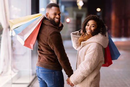 Happy Loving Family. Portrait of smiling black man and woman turning back and looking at camera over the shoulder holding hands, carrying shopping bags. Cheerful african american couple near mallの写真素材