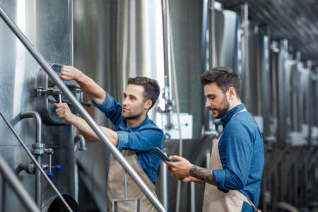 Modern devices for work at brewery. Millennial guy worker in apron turns a valve on big boiler for lager, manager with tablet controls process in interior of plant with equipment, free spaceの写真素材