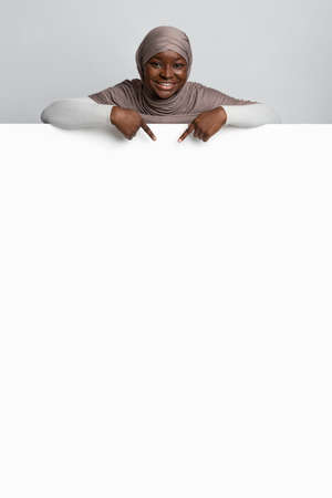 Look There. Cheerful Black Muslim Lady In Hijab Pointing With Fingers At Blank White Advertisement Board, Indicating Copy Space For Your Text Or Design, Standing Over Light Background, Vertical Shotの写真素材