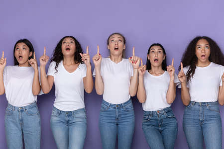 Five Amazed Multiracial Ladies Pointing Fingers Up Standing Together Posing Over Purple Studio Background. Look Upward, Check This Out Concept, Advertisement Bannerの写真素材