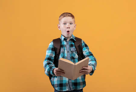 Shocked schooler reading book on yellow studio background. Redhead schoolboy with backpack holding...
