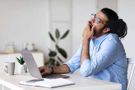 Sleepy Freelancer Guy Yawning At Workplace In Home Office, Tired Of Work With Laptop, Tired Western Man Sitting At Desk With Computer And Covering Mouth With Hand, Free Spaceの写真素材