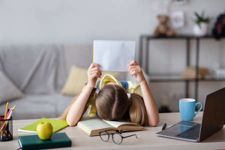 Teenage girl sitting at table, resting head on paper book, holding piece of paper, tired of school,...