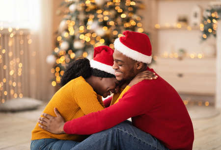 Free time together and new year at home during self-isolation. Laughing young african american guy and lady in Santa Claus hats hugging in living room with Xmas tree and decor with glowing garlandsの写真素材
