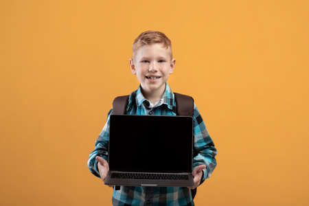 Cheerful ginger kid with schoolbag showing laptop with blank screen on yellow studio background, copy space. Happy schoolboy holding modern laptop, using technologies in education, mockupの写真素材