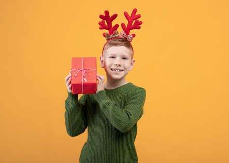 Happy kid with New Year gift over yellow studio background, copy space. Excited boy with deer accessory holding xmas present next to ear, wondering what is inside. Winter holidays celebrationの写真素材